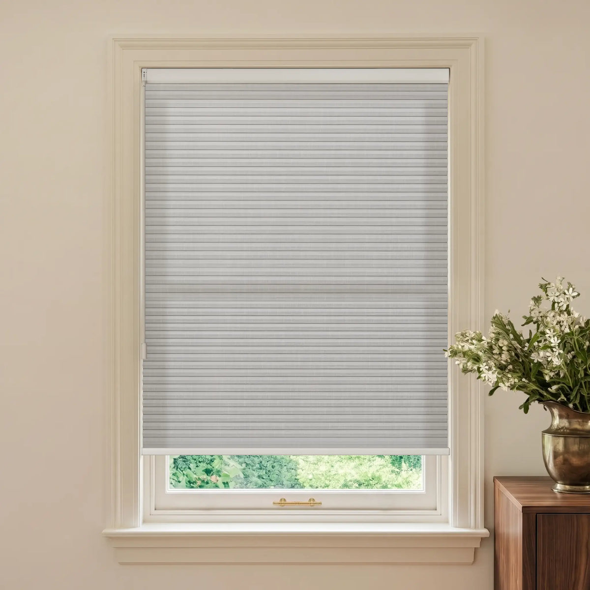 A light gray cellular window shade installed in a white window frame, shown in a styled room setting with a wooden side table and a vase of white flowers on the right.
