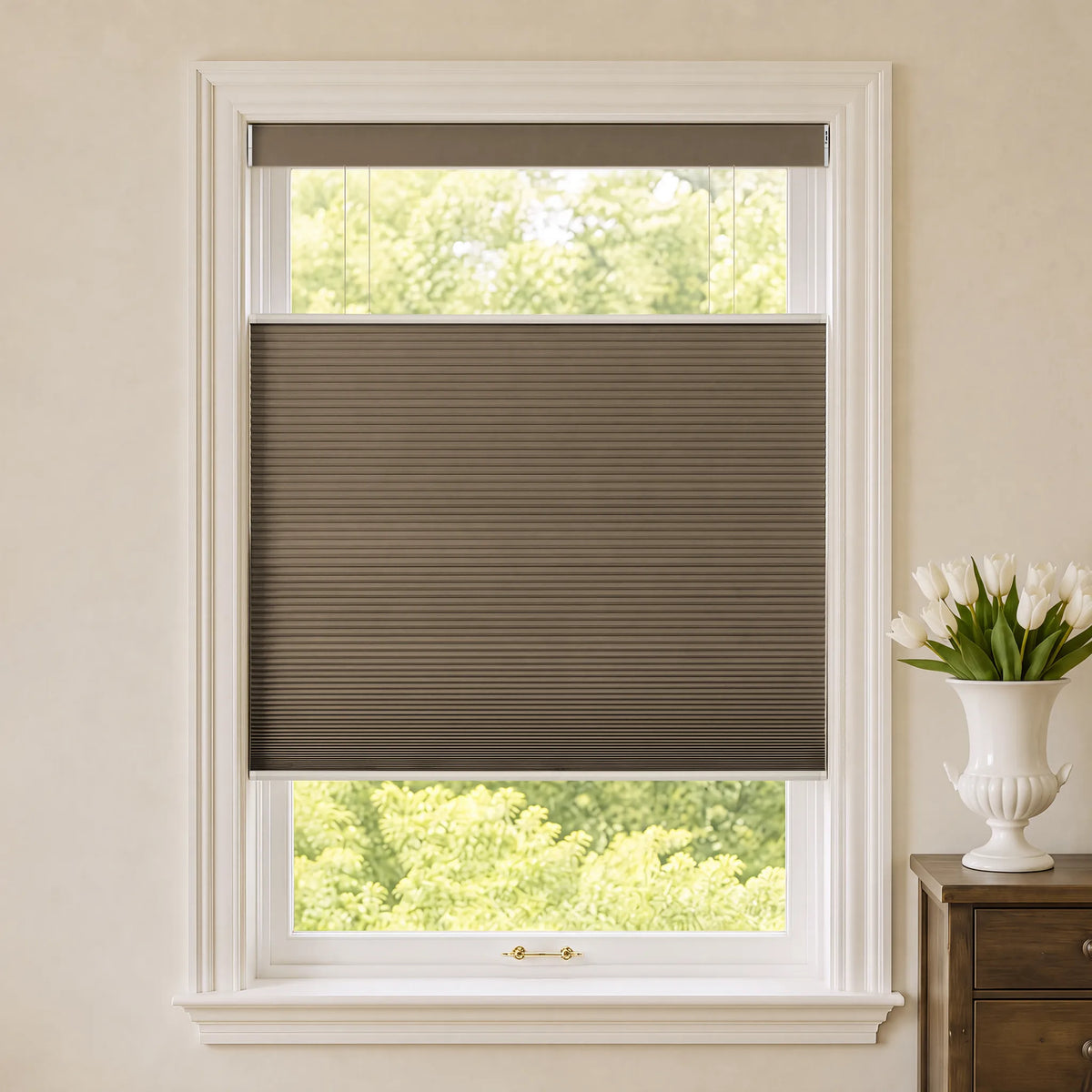 A brown Motorized Top Down Bottom Up Blackout Cellular Shades installed on a white-framed window, with a view of green trees outside and a white vase of tulips on a wooden side table to the right.