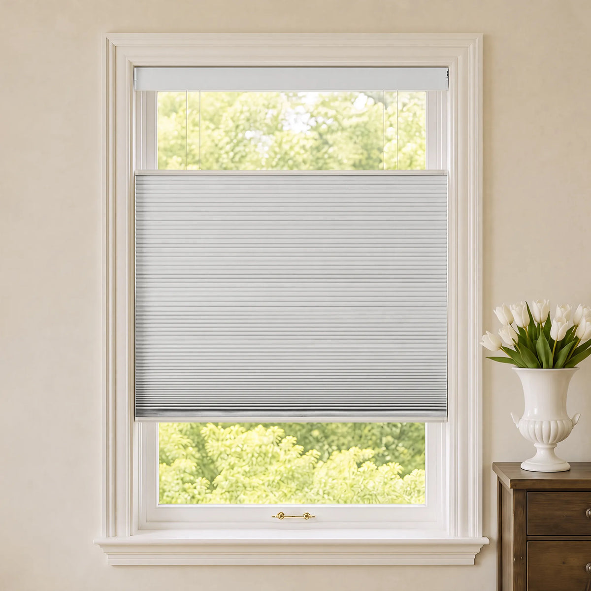 A white Motorized Top Down Bottom Up Blackout Cellular Shades partially drawn over a white-framed window, with a view of green trees outside and a white vase of tulips on a wooden side table to the right.