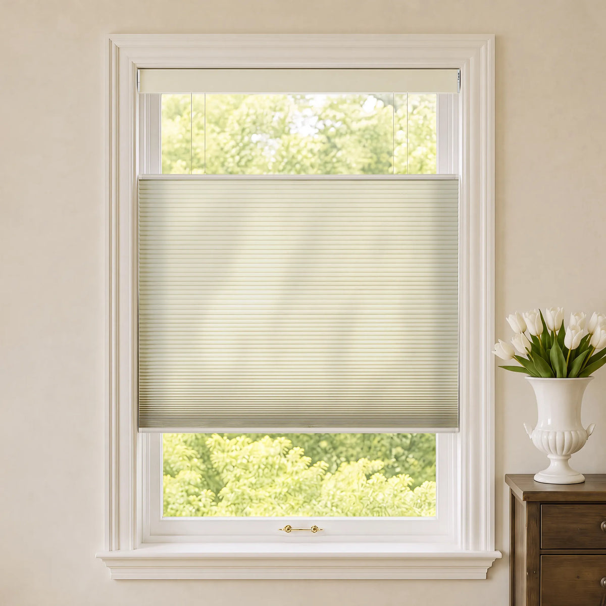 A beige Motorized Top Down Bottom Up Cellular Shades window blind shown installed on a white-framed window in a bright room, with a vase of white tulips on a wooden side table to the right.