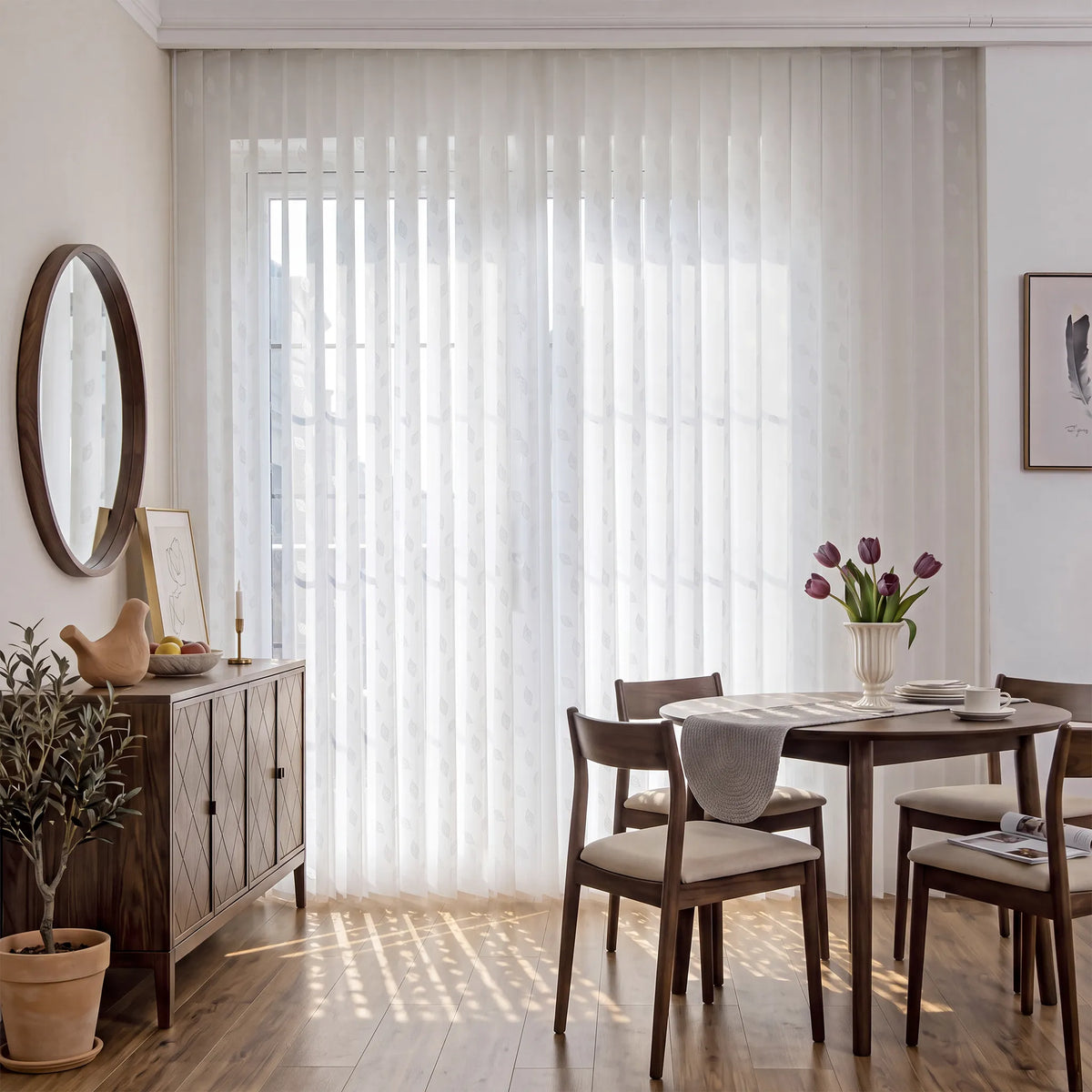 A dining room scene with white Smart Sheer Vertical Blinds covering a large window, styled with a wooden sideboard, chairs, a round mirror, and a vase of tulips on the table.