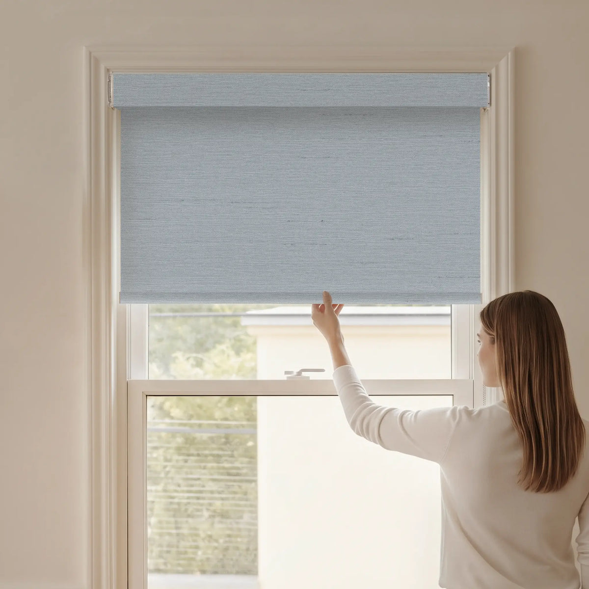 Woman adjusting a Graywind blackout manual-motorized roller shade in Horizon (Sky Blue) fabric.
