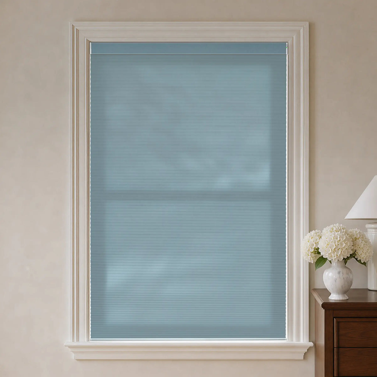 A light blue-gray cellular window shade installed in a white-trimmed window, shown in a styled room setting with a wooden side table, white flowers, and a lamp on the right.
