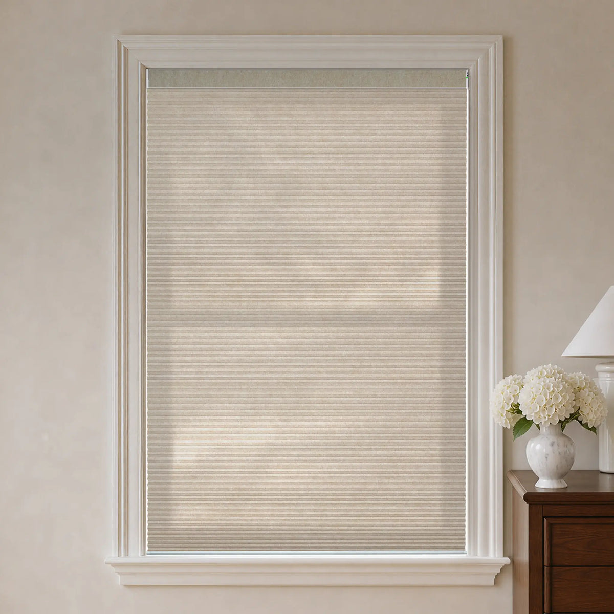 Beige cellular window shade installed in a white-framed window, shown in a styled room setting with a wooden side table and a vase of white flowers on the right.