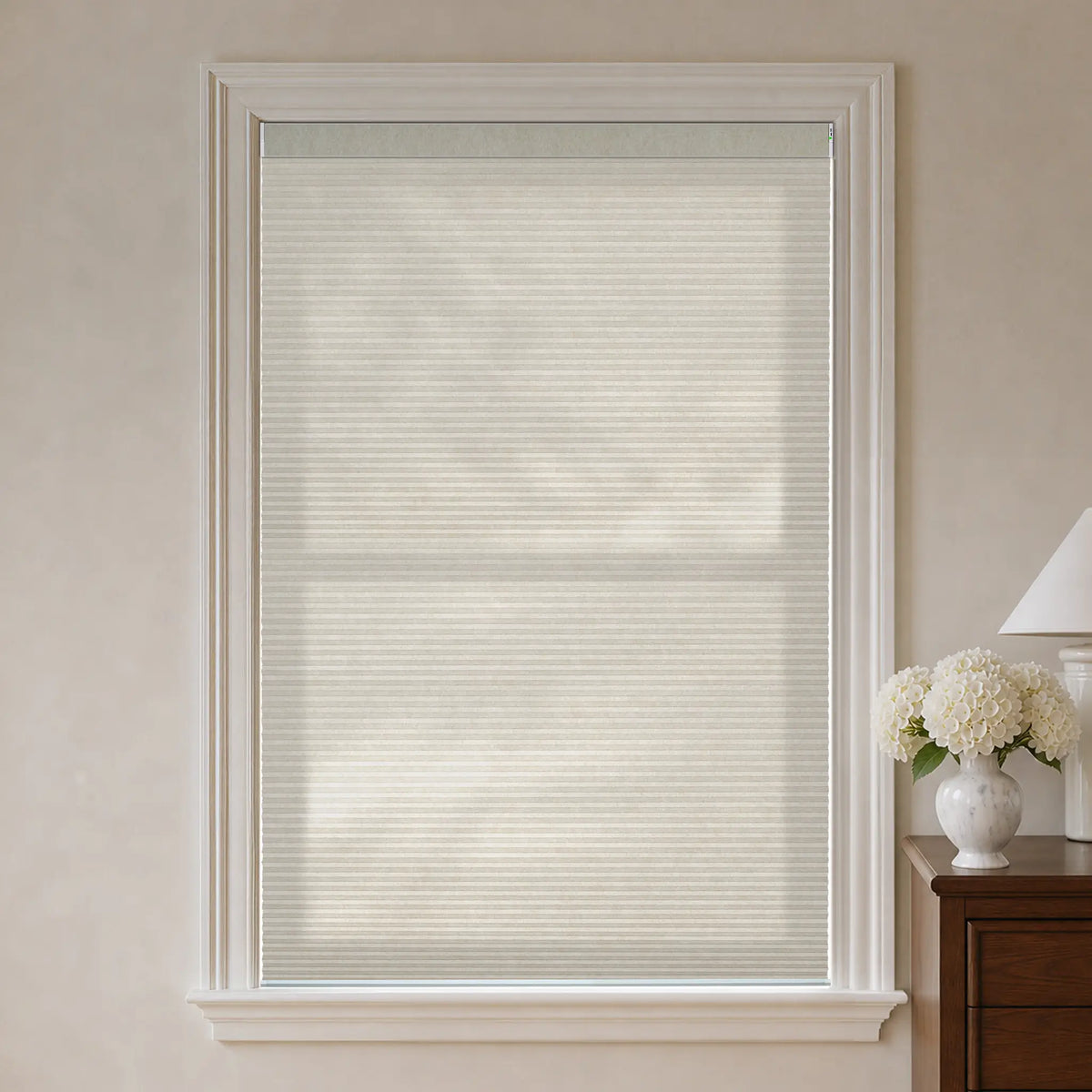 A beige cellular window shade installed in a white-framed window, shown in a styled room setting with a wooden side table, lamp, and white flowers on the right.