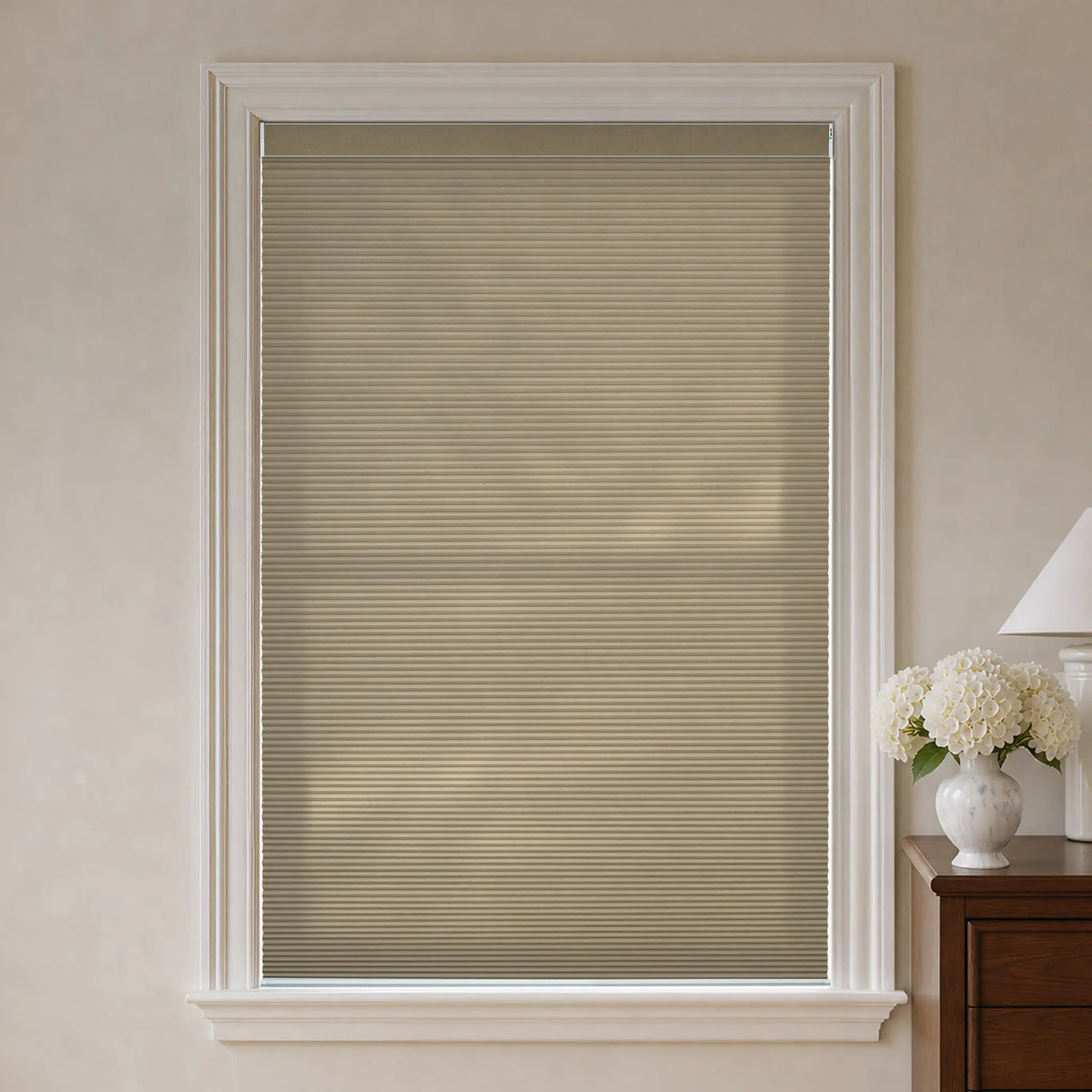 Beige cellular window shade installed in a white window frame, shown in a styled room setting with a wooden side table, white flowers, and a lamp on the right.