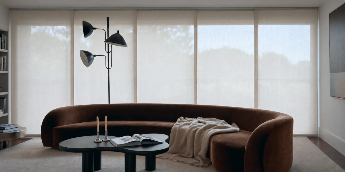 A modern curved brown sofa in a bright, contemporary living room with large windows covered by sheer white roller blinds. A black multi-arm floor lamp, a round black coffee table with candles and an open book, and a beige throw blanket are arranged in front of the sofa.