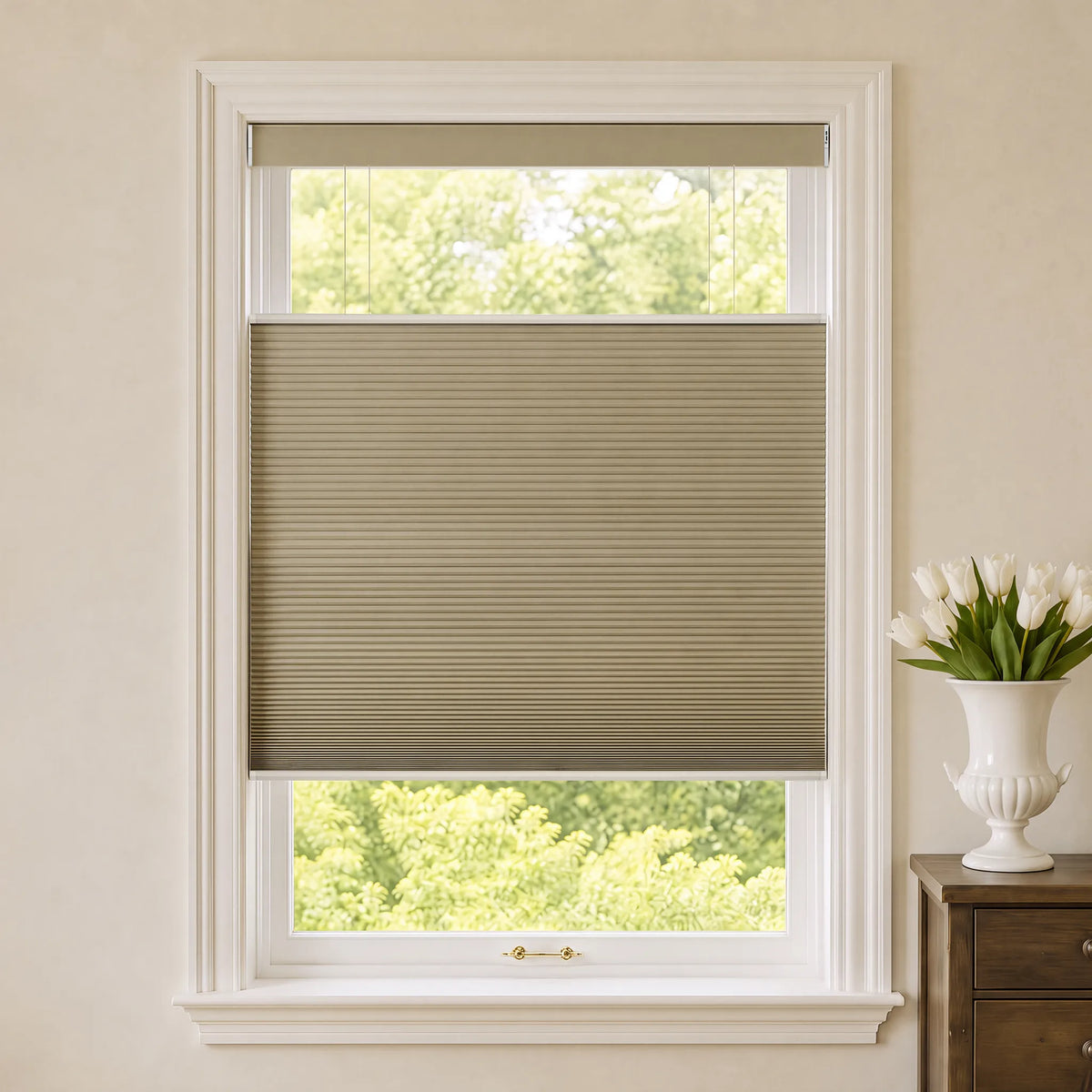 A beige A sheer white Motorized Top Down Bottom Up Cellular Shades  partially drawn over a white-framed window, with a view of green trees outside and a white vase of tulips on a wooden side table to the right.e with a top roller panel, shown installed on a white-framed window in a bright room with a vase of white tulips on a wooden side table.