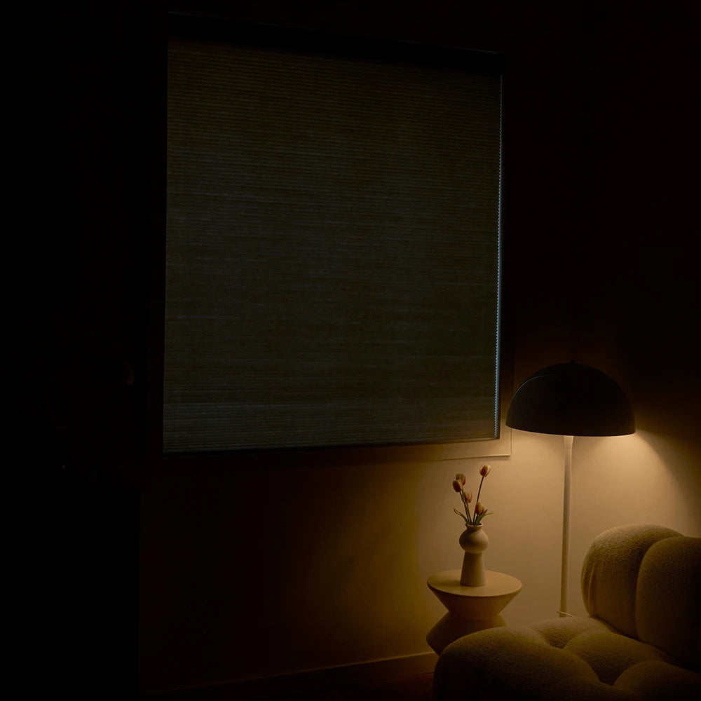 Dark room with cordless top down bottom up cellular shades​, a lamp casting light on a wall and a vase with flowers on a small table.
