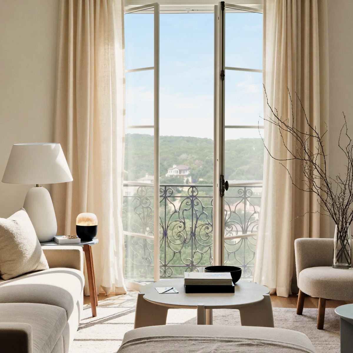 Modern living room with large windows, beige sheer curtains, and a view of greenery outside.
