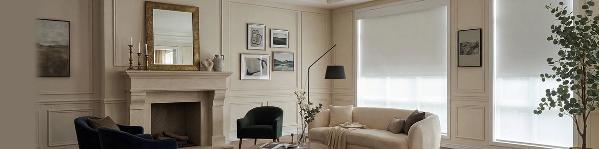 A styled living room interior with cream walls, a fireplace mantel, framed wall art, a beige sofa, dark accent chairs, a floor lamp, and large windows with white dual shades.
