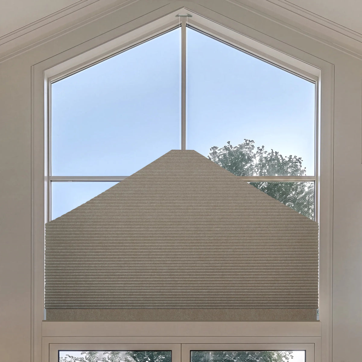 A coffee Elongated Triangle Cellular Shades installed on a large arched window, with a view of blue sky and trees outside.