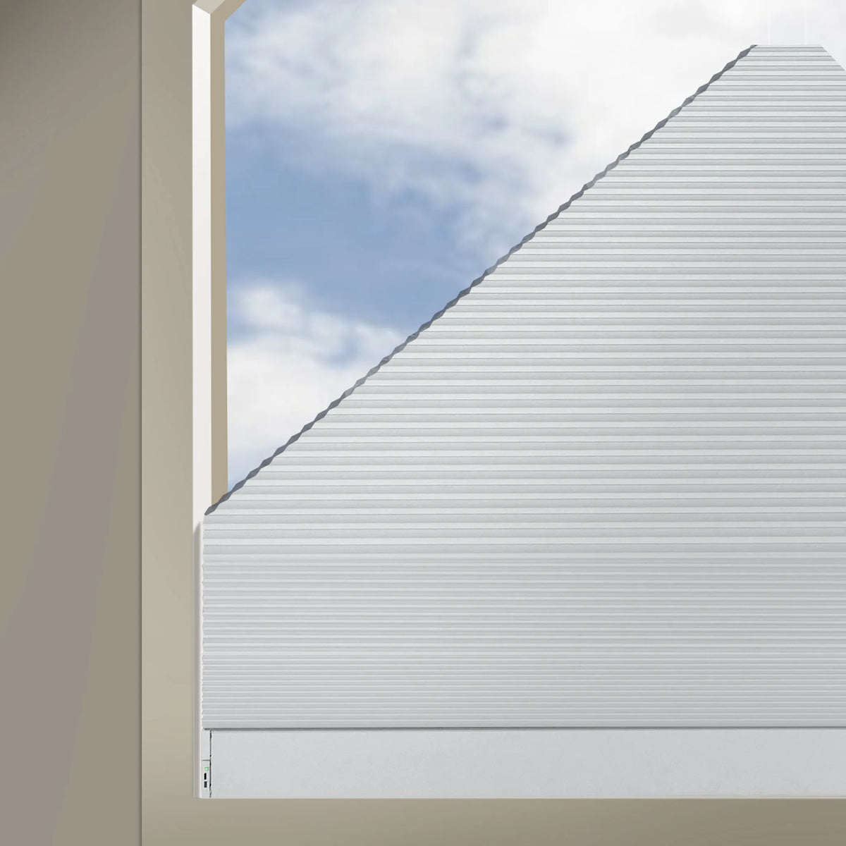 A close-up of a white Elongated Triangle Cellular Shades, shown partially drawn across a window opening with beige walls and a blue sky with clouds visible outside.
