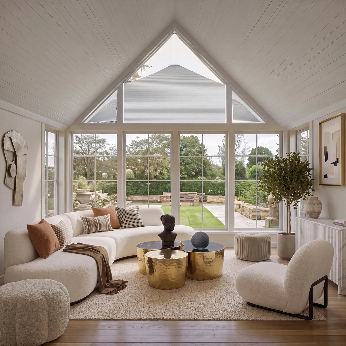 A bright, modern living room with a curved cream sofa, textured poufs, gold metallic coffee tables, and a large window looking out onto a landscaped garden.