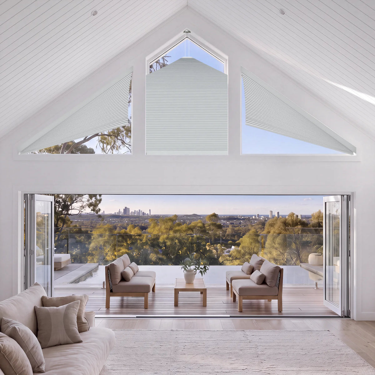 A bright, modern living room with a high white-painted wood ceiling, large arched windows, and open glass doors revealing a deck with outdoor seating and a distant city and forest view.