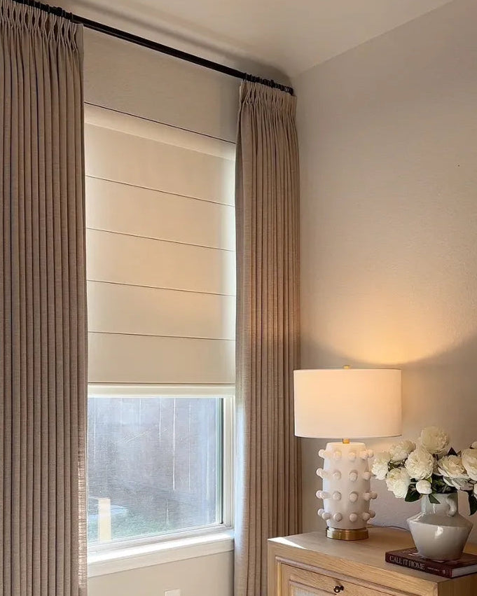 A cozy bedroom corner showing a window with a beige Roman shade paired with matching pleated drapes and a decorative table lamp.