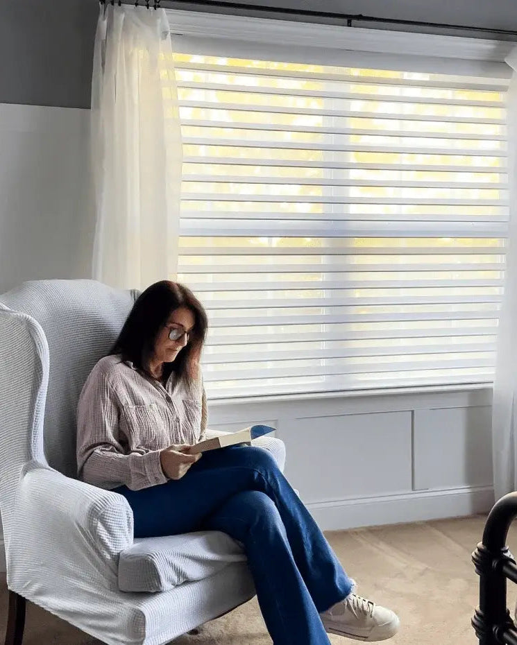 A woman reading a book in a grey wingback chair next to a large window with white horizontal blinds and sheer side panels.