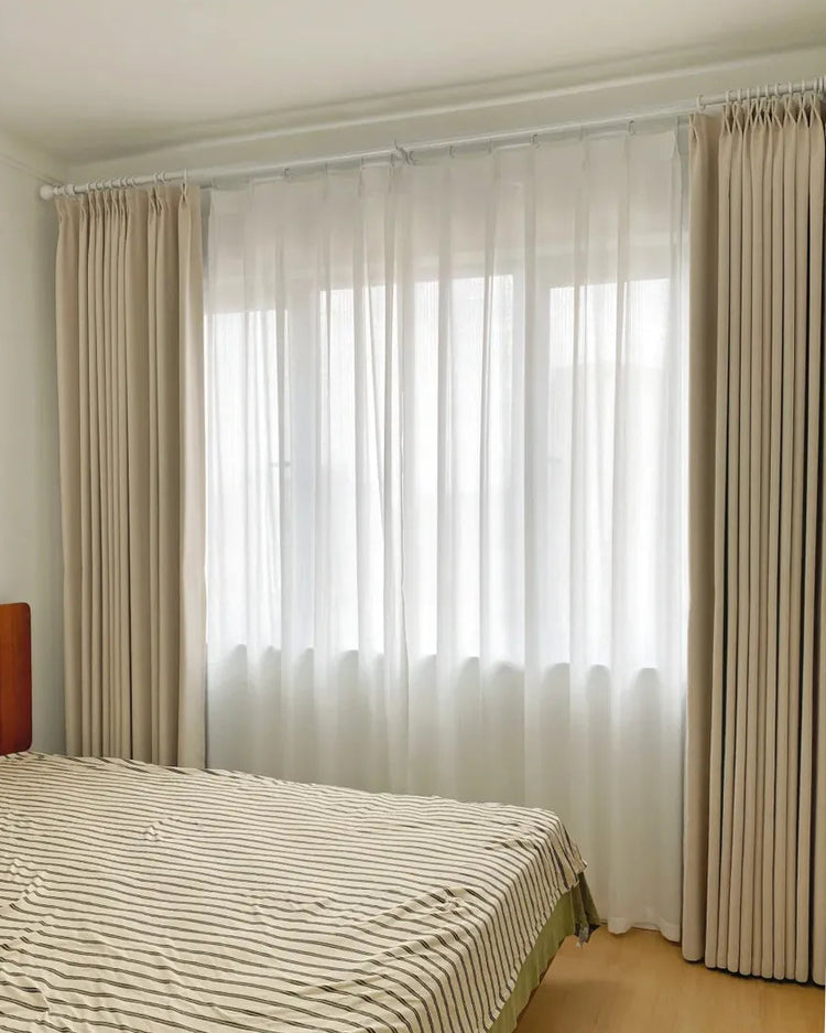 A bedroom window setup with a double-layer window treatment consisting of light-filtering white sheers and beige blackout drapes.