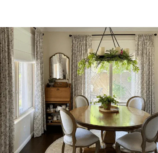 A classic dining room with a round wooden table, a greenery-adorned chandelier, and windows decorated with floral patterns and Roman shades.