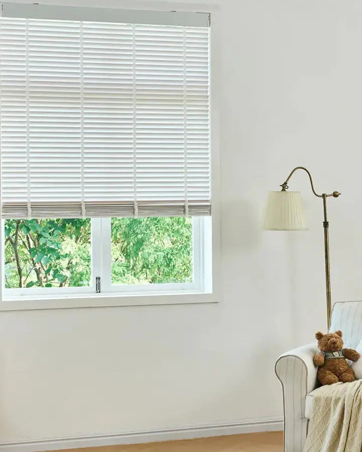 White minimalist living room featuring a window with white wood horizontal blinds, a floor lamp, and a teddy bear on an armchair.