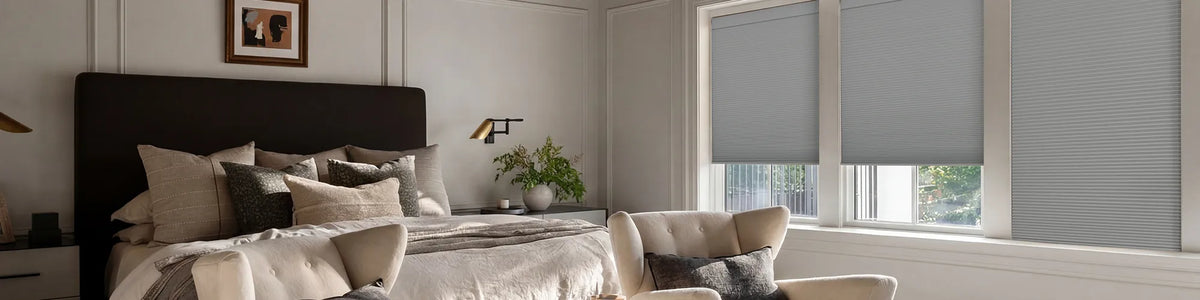 A bedroom interior showing gray cellular window blinds installed on two windows, with a bed, pillows, chair, and wall decor visible in the room.