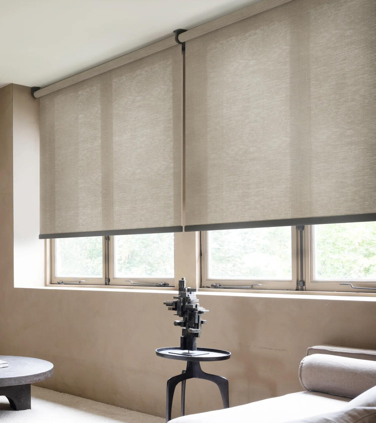Beige roller blinds covering a window in a room with a chair and small table.