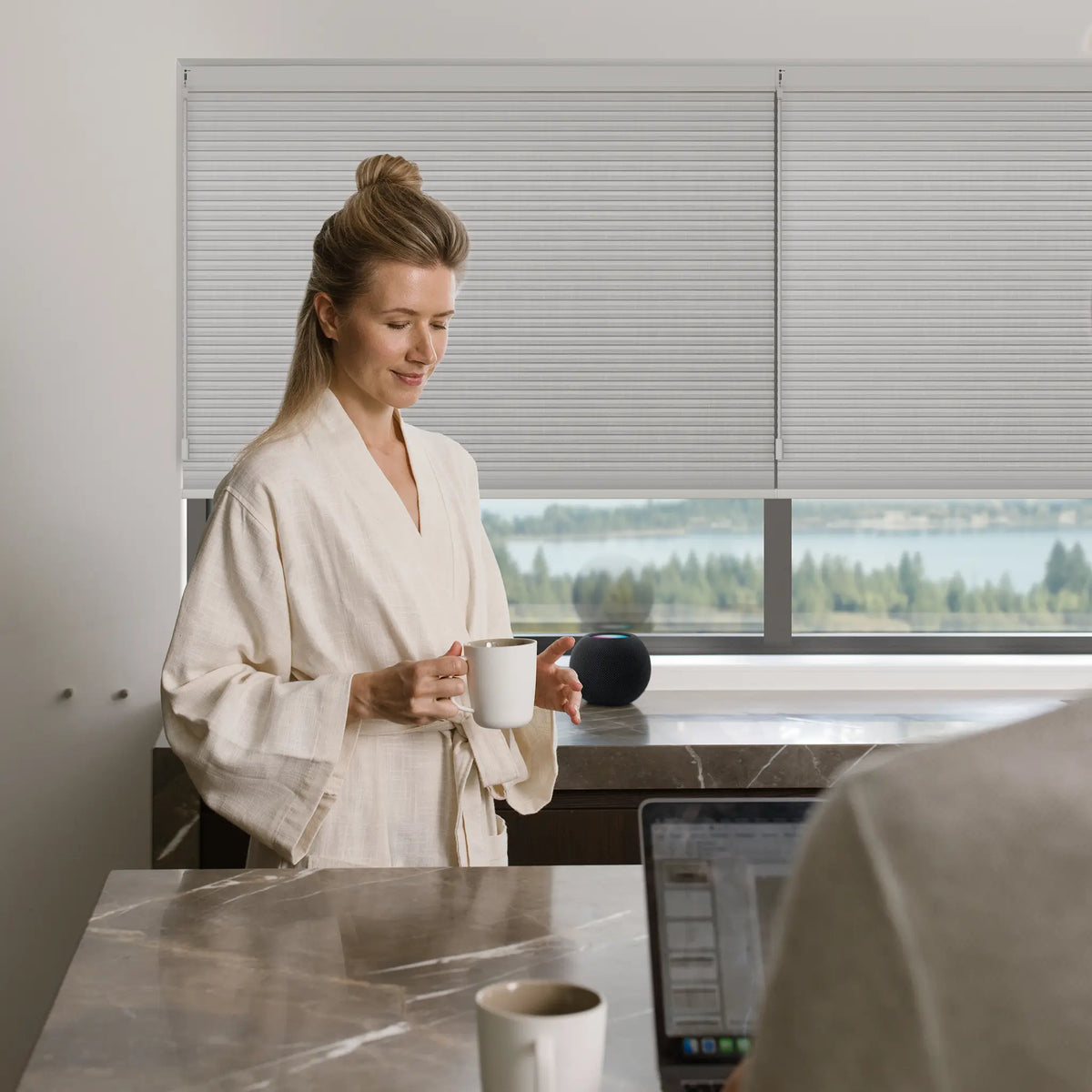 A woman in a cream linen robe stands by a window holding a white mug, with a modern interior, marble countertop, and outdoor view in the background.