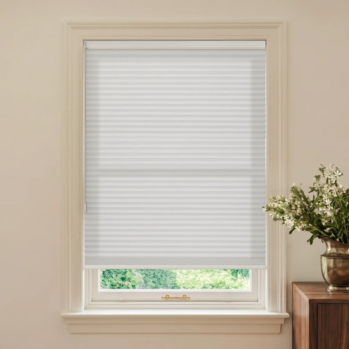 White hMotionblinds Smart Cellular Shades  installed on a window in a beige room, with a wooden side table and a vase of white flowers visible on the right.