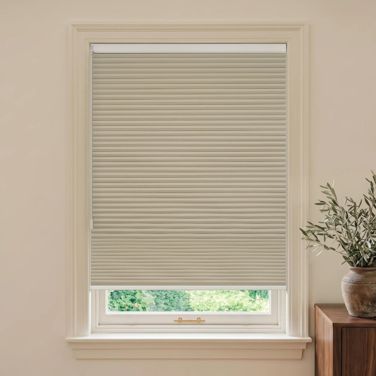 A beige Motionblinds Smart Cellular Shades  partially drawn over a white-framed window, shown installed in a neutral cream-colored room with a wooden side table and potted olive branches visible on the right.