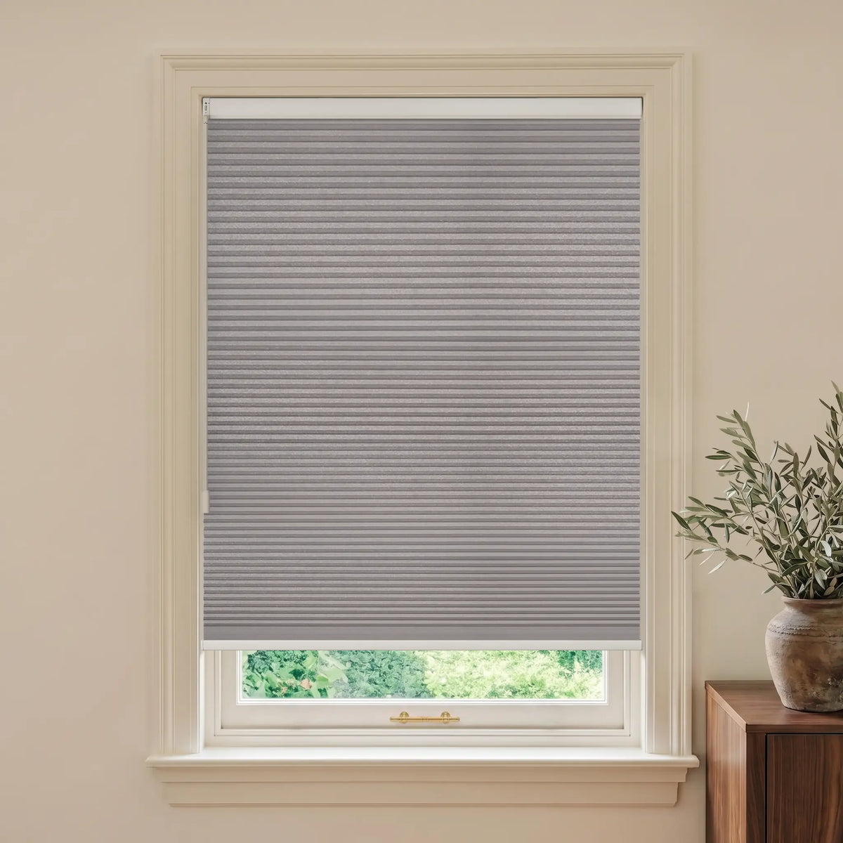A gray cellular window shade installed in a white window frame, shown in a styled room setting with a potted olive branch on a wooden side table.