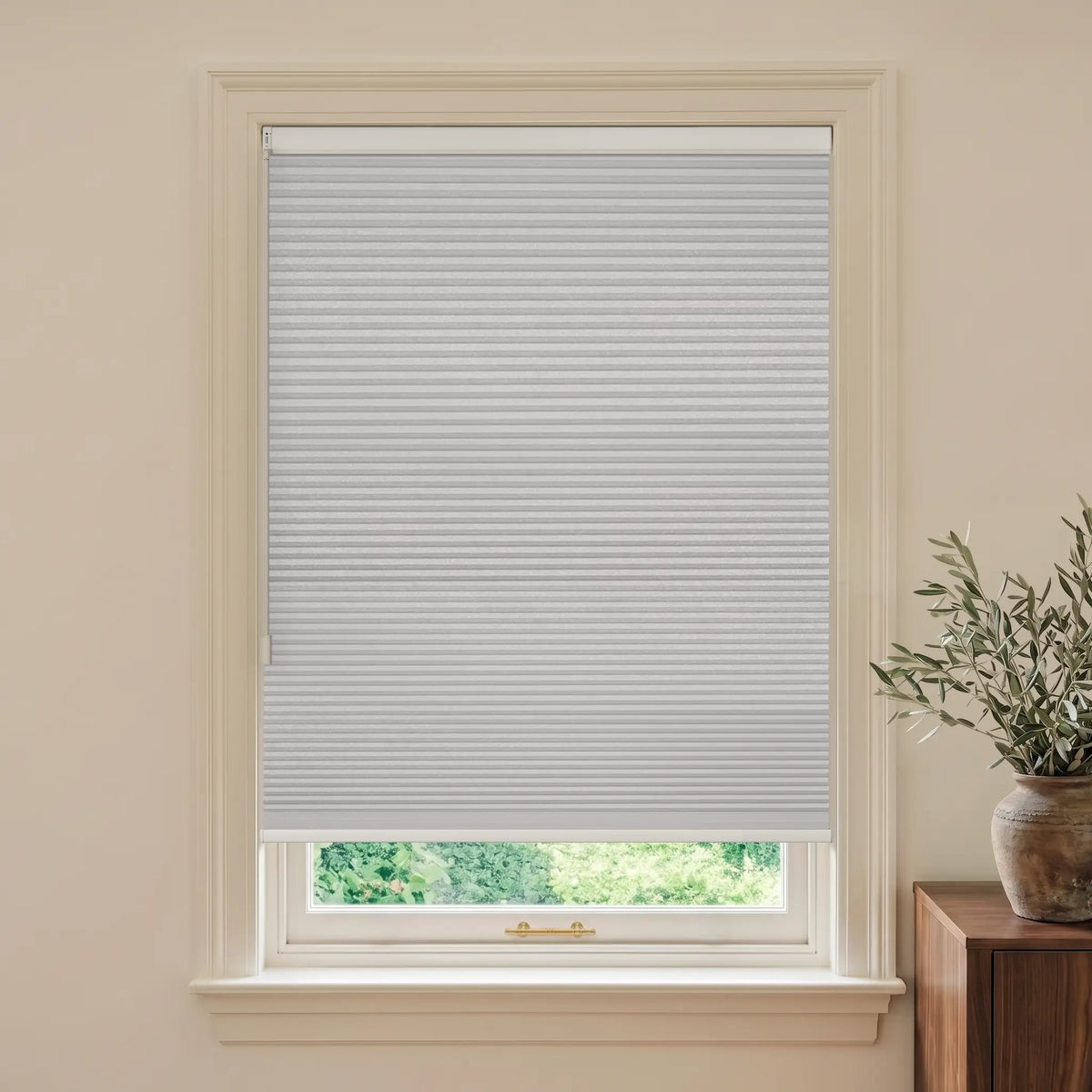 A white cellular window shade installed in a white window frame, shown in a styled room setting with a beige wall and a potted plant on a wooden side table.