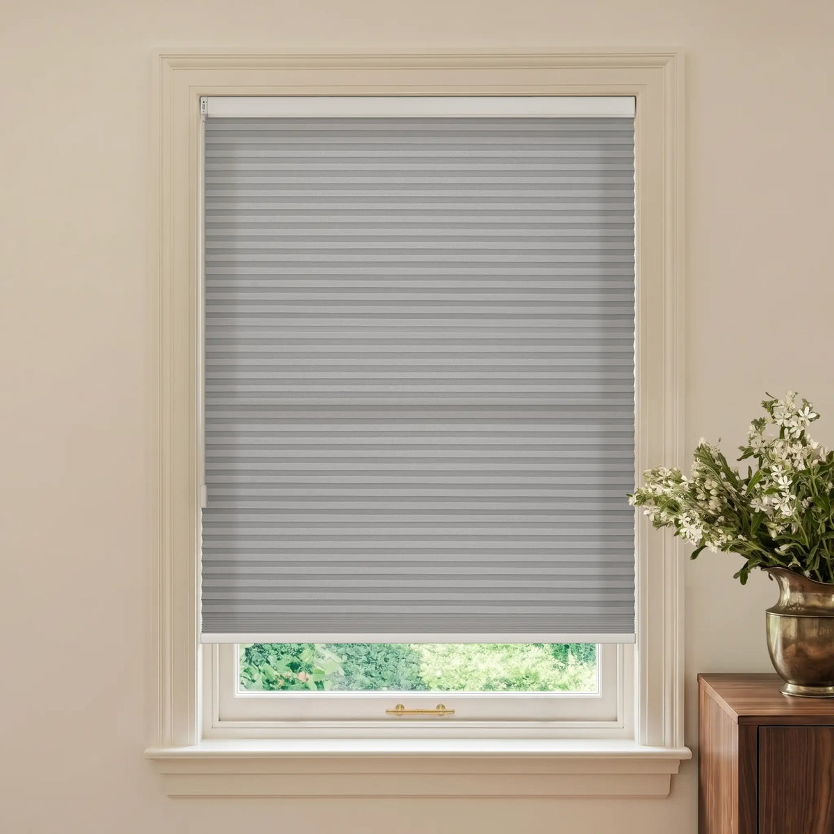 A gray cellular window shade installed in a white window frame, shown in a styled room setting with a beige wall and a wooden side table holding a vase of white flowers.