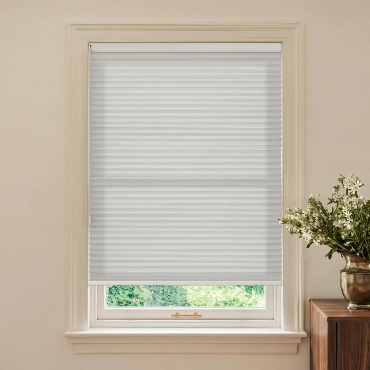 A white cellular window shade installed in a white window frame, shown in a styled room setting with a beige wall and a wooden side table holding a vase of white flowers.