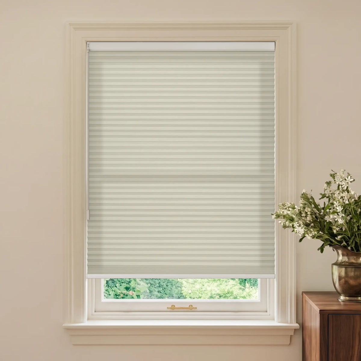 A window with a partially drawn beige cellular shade, set in a cream-colored wall with a wooden side table and a vase of white flowers visible on the right.