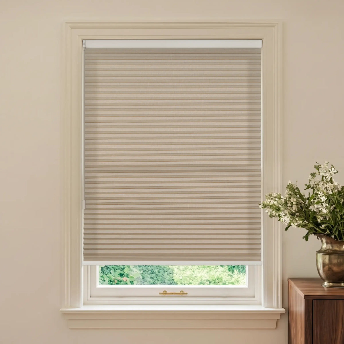 A coffee cellular window shade installed in a white window frame, shown in a styled room setting with a wooden side table and a vase of white flowers on the right.
