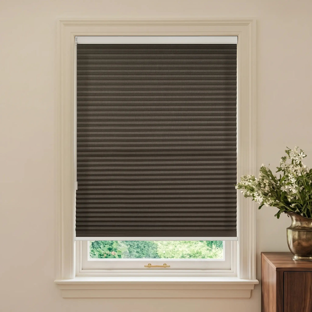 A dark brown Motionblinds Smart Cellular Shades installed in a white window frame, shown in a styled room setting with a wooden side table and a vase of white flowers on the right.