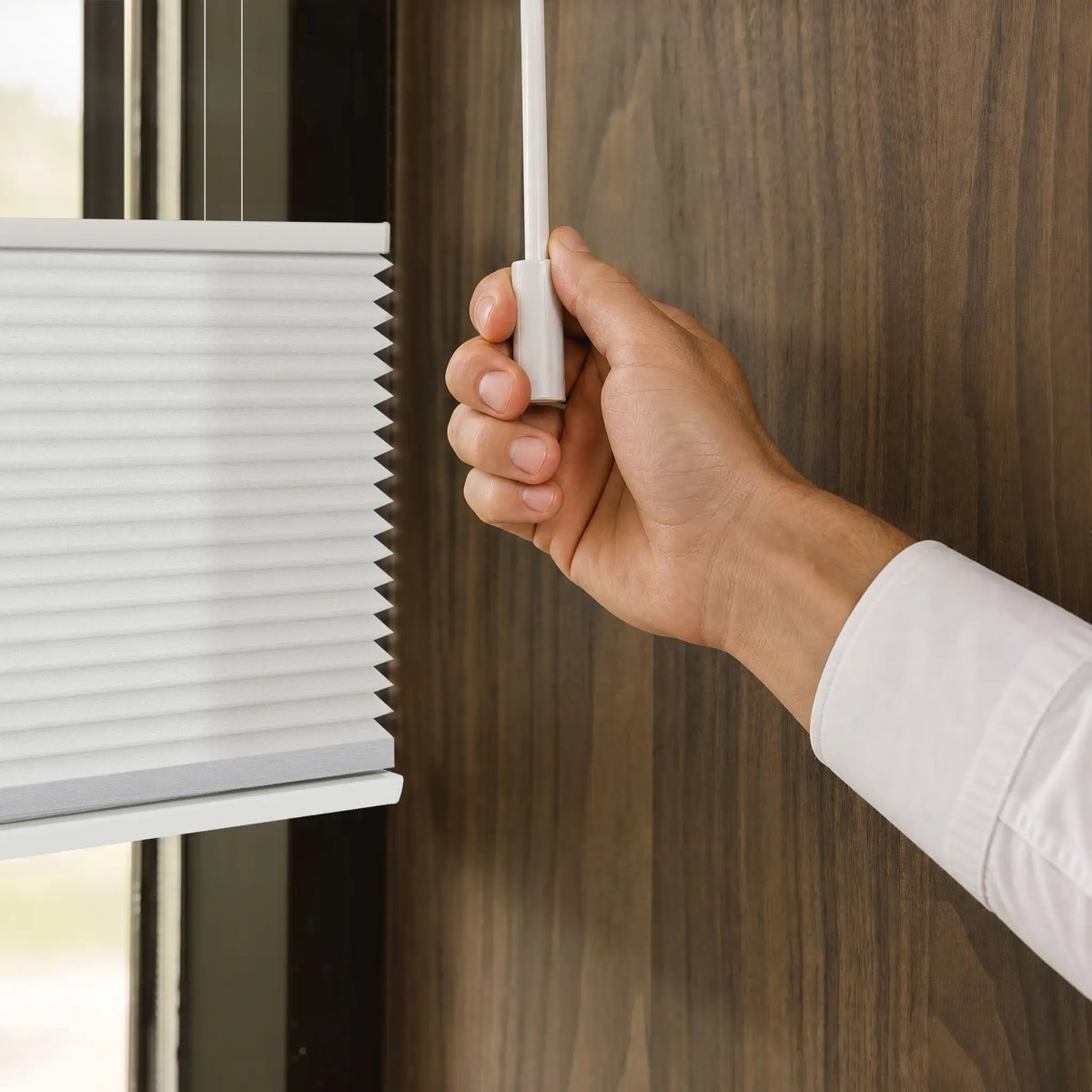 A close-up of a hand pulling a wand to operate a white Motionblinds Smart Top Down Bottom Up Cellular Shades against a dark wood-paneled wall.