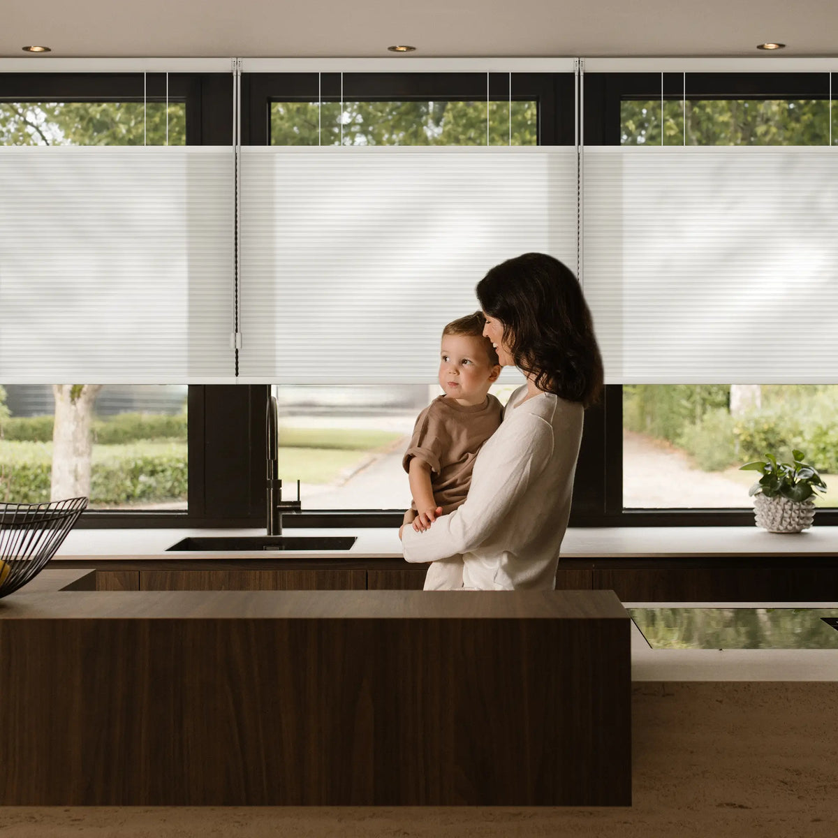 A woman holding a child in a modern kitchen, with whiteMotionblinds Smart Top Down Bottom Up Cellular Shades partially drawn on large windows behind them.