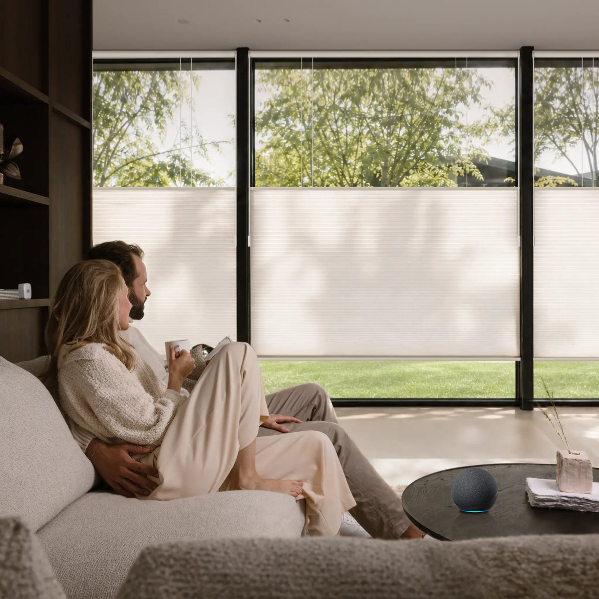 A couple sits on a light beige sofa in a modern living room, looking out large black-framed windows with Motionblinds Smart Top Down Bottom Up Cellular Shades . The room is brightly lit with natural daylight, showing green trees and grass outside.