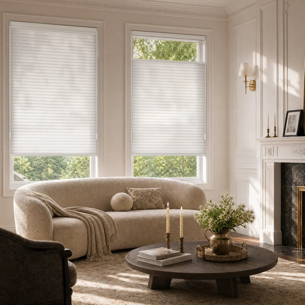 A styled living room with two white Motionblinds Smart Top Down Bottom Up Cellular Shades  on tall windows, a beige curved sofa, a round dark wood coffee table, and a fireplace in the background.