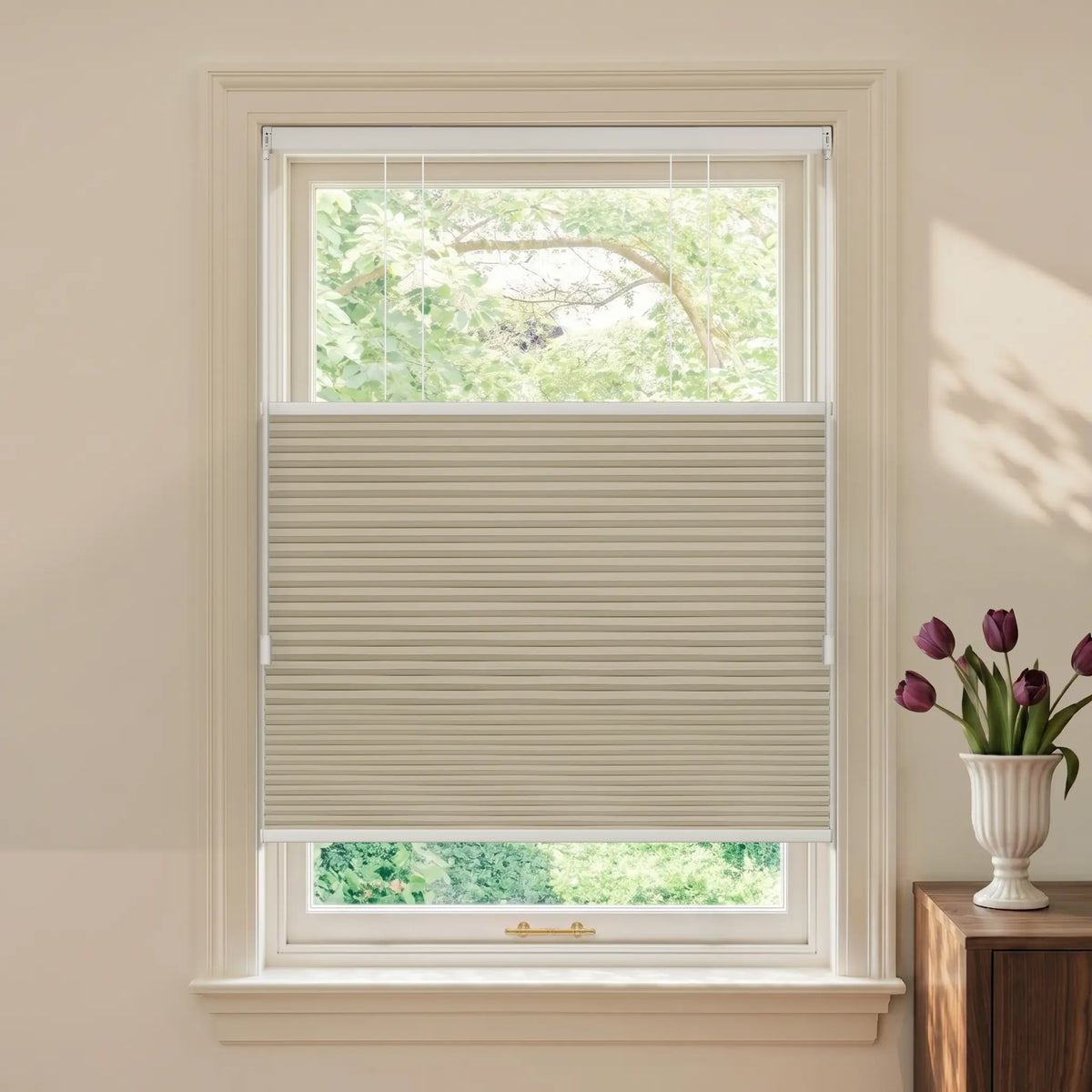A beige Motionblinds Smart Top Down Bottom Up Cellular Shades partially drawn across a white-framed window, shown in a styled room setting with a vase of purple tulips on a wooden side table.
