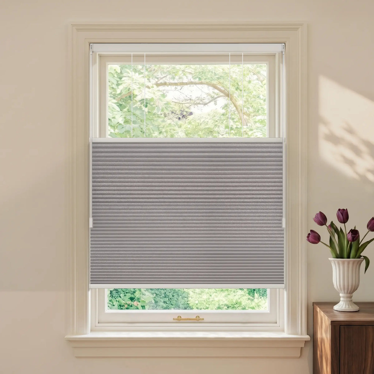 A gray Motionblinds Smart Top Down Bottom Up Cellular Shades  partially drawn across a white-framed window, shown in a styled room setting with a vase of tulips on a wooden side table.