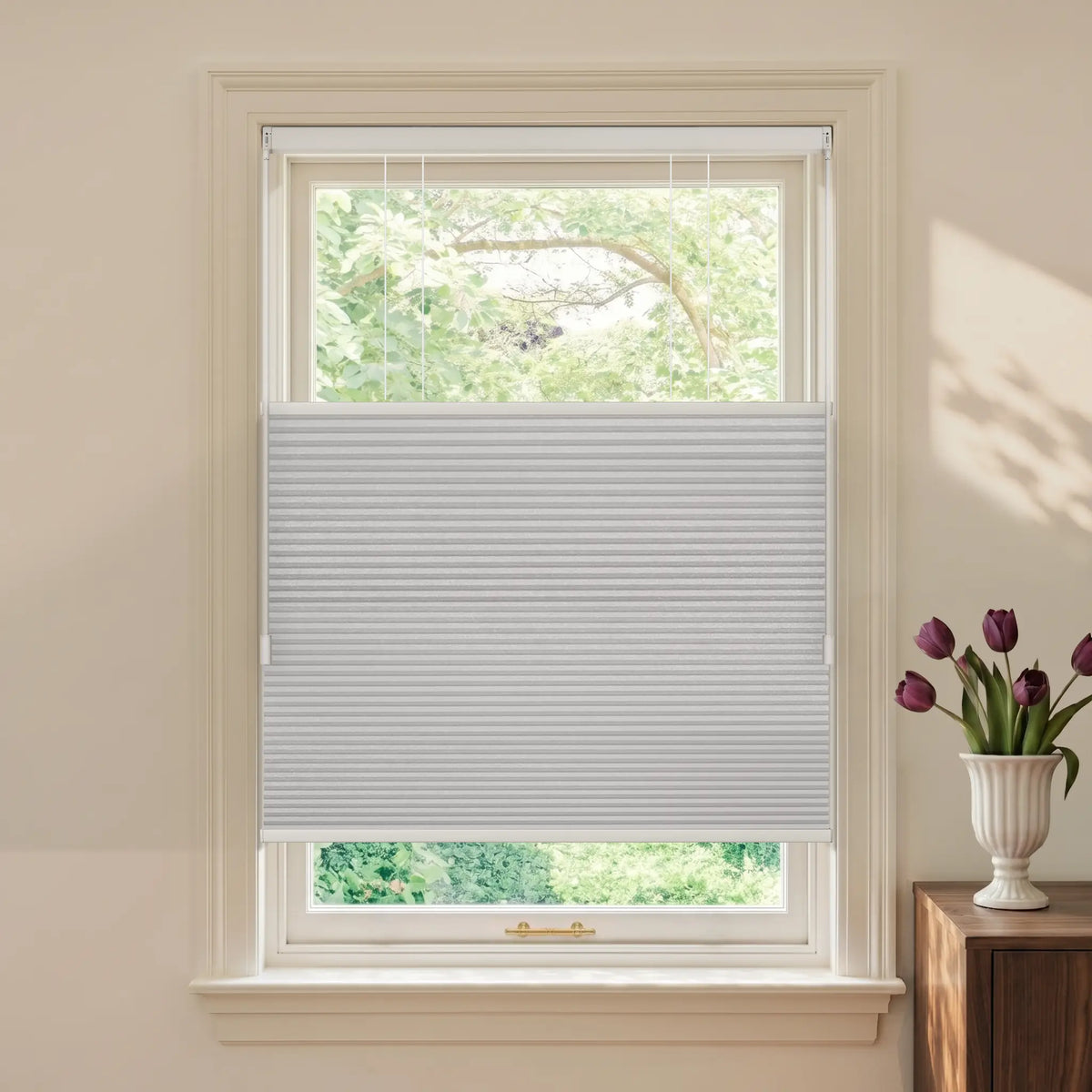 A white Motionblinds Smart Top Down Bottom Up Cellular Shades  partially drawn across a white-framed window, shown in a styled room setting with a vase of purple tulips on a wooden side table.