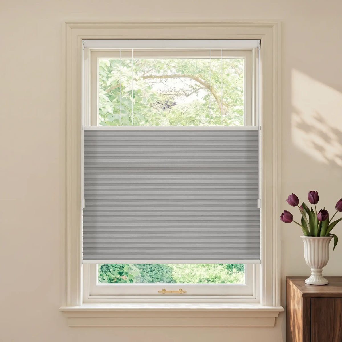 A gray Motionblinds Smart Top Down Bottom Up Cellular Shades installed in a white window, shown in a styled room with a cream wall, a wooden side table, and a vase of purple tulips.