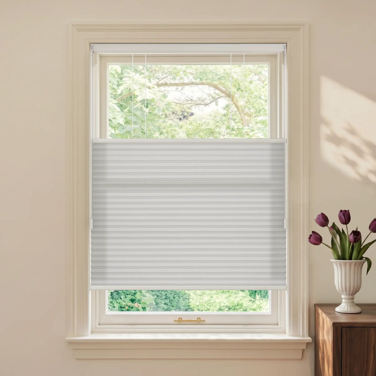 A Motionblinds Smart Top Down Bottom Up Cellular Shades  partially drawn across a white-framed window, shown in a styled room setting with a vase of purple tulips on a wooden side table.