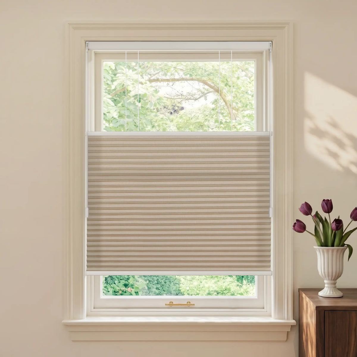 A beige Motionblinds Smart Top Down Bottom Up Cellular Shades  installed in a white window frame, shown in a styled room setting with a vase of purple tulips on a wooden side table.