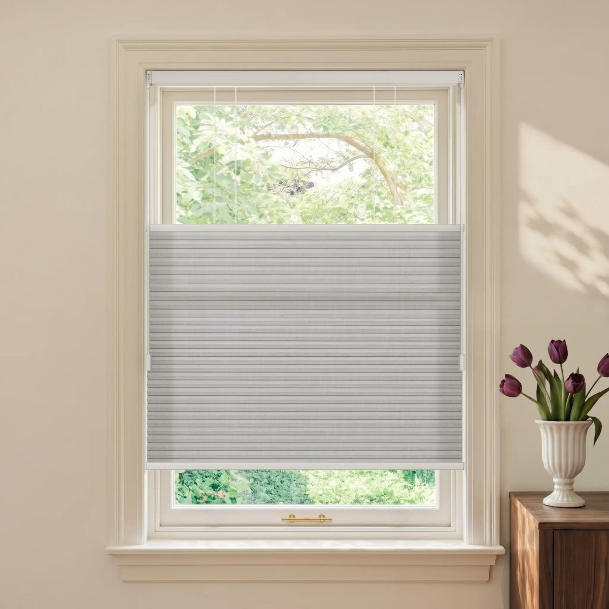 A light gray Motionblinds Smart Top Down Bottom Up Cellular Shades installed in a white window, shown in a styled room with a cream wall, a wooden side table, and a white vase of purple tulips.