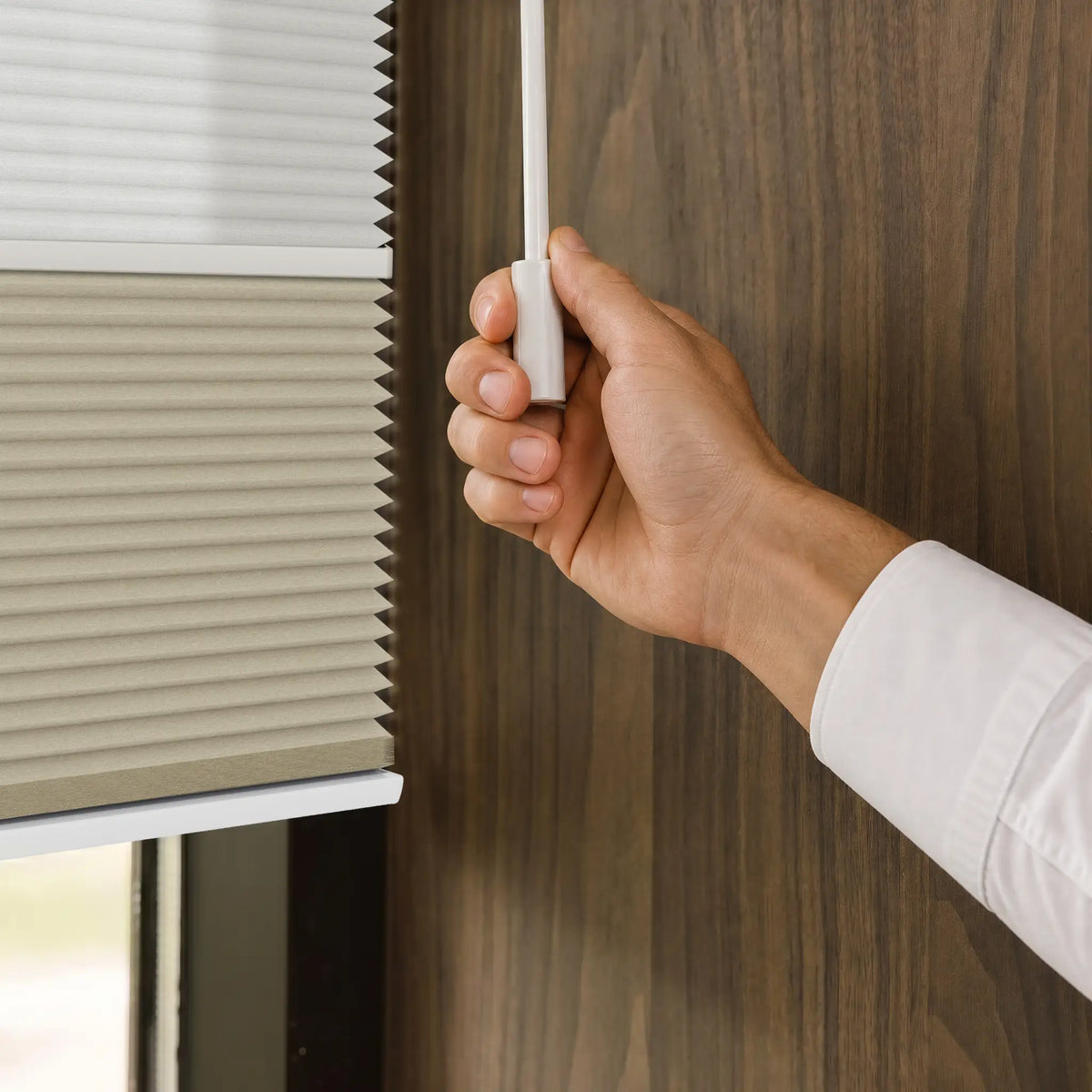 A close-up of a person's hand holding a white pull tab to operate a Motionblinds smart Day Night Cellular Shades  next to a dark wood-paneled wall.