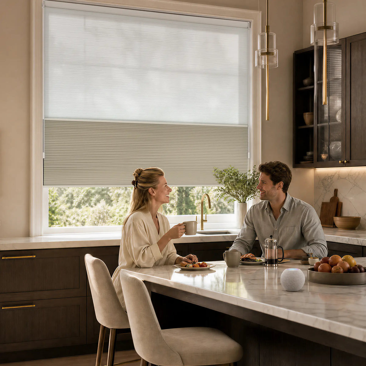 A modern kitchen scene with a couple seated at a marble island, with a large window above them featuring a partially drawn Motionblinds smart Day Night Cellular Shades . The room has warm beige walls, dark wood cabinetry, pendant lights, and a fruit bowl on the counter.