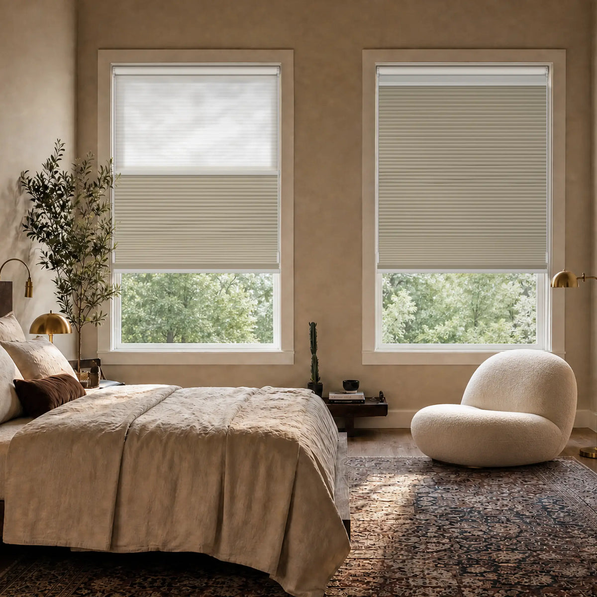 A styled bedroom interior showing two Motionblinds smart Day Night Cellular Shades  partially drawn. The room includes a bed with neutral bedding, a sculptural white chair, lamps, a rug, and visible greenery outside the windows.